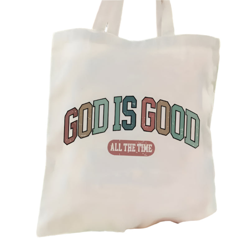 Tote bag with 'God is Good All the Time' text held against a green outdoor background