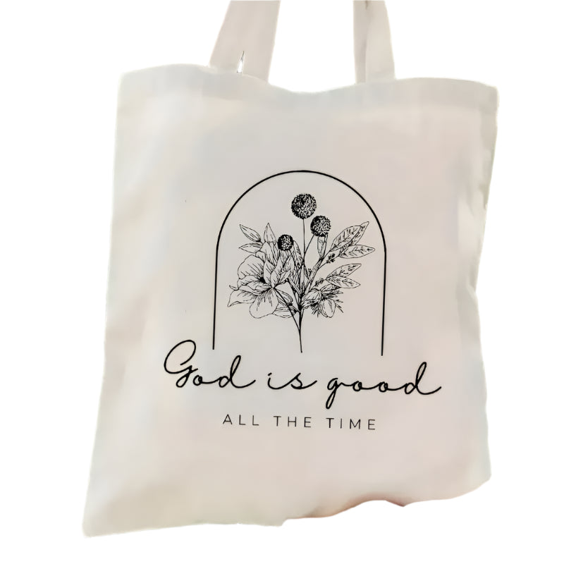 Tote bag with floral design and 'God is good all the time' text held in front of green foliage.