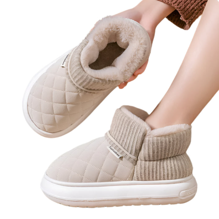 Beige winter boots with white soles being held by a hand on a wooden floor.