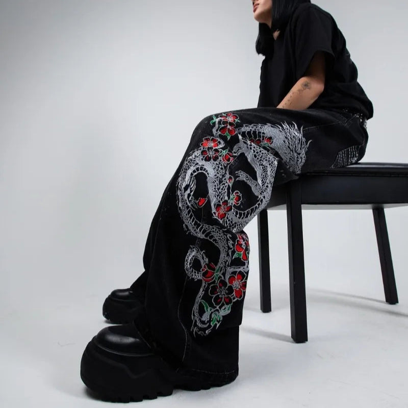 Person wearing black pants with floral embroidery sitting on a chair against a white background
