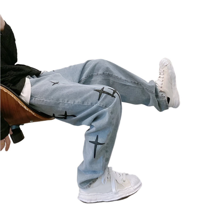 Person wearing light blue jeans with black cross designs and white sneakers, sitting on a chair.