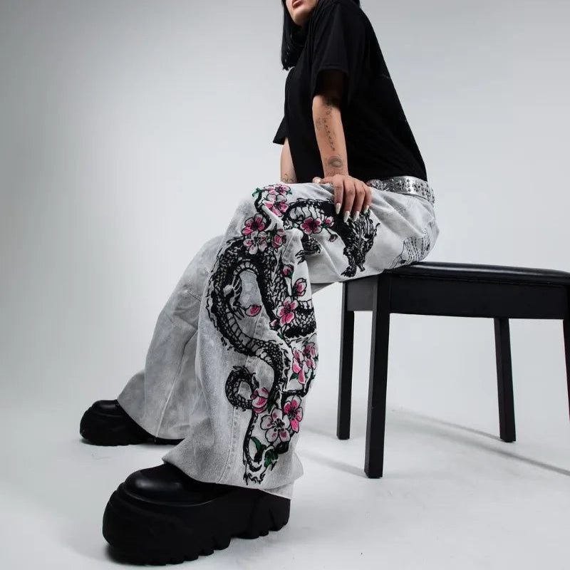 Person wearing a black top and white pants with floral patterns, sitting on a chair against a gray background.
