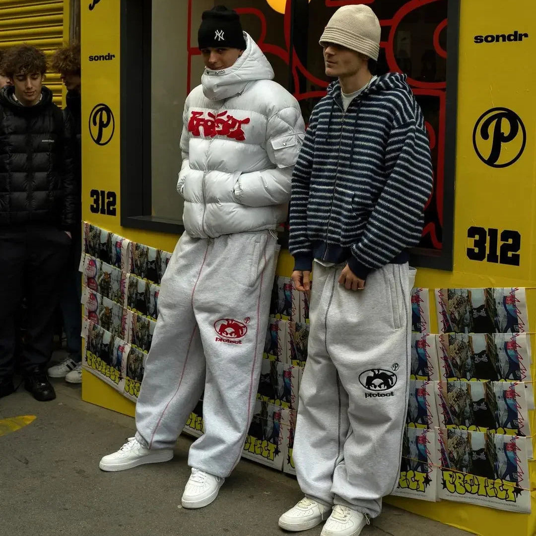 Two people wearing matching white tracksuits with red and black logos in front of a yellow store.