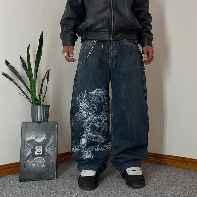 Person wearing a black jacket and blue jeans with a dragon design, standing in a room with a plant and a box.