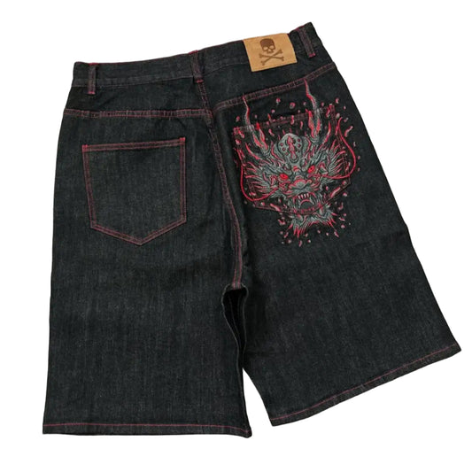 Black denim shorts with a colorful embroidered design on a gray concrete surface