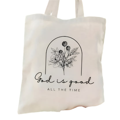 Tote bag with floral design and 'God is good all the time' text held in front of green foliage.