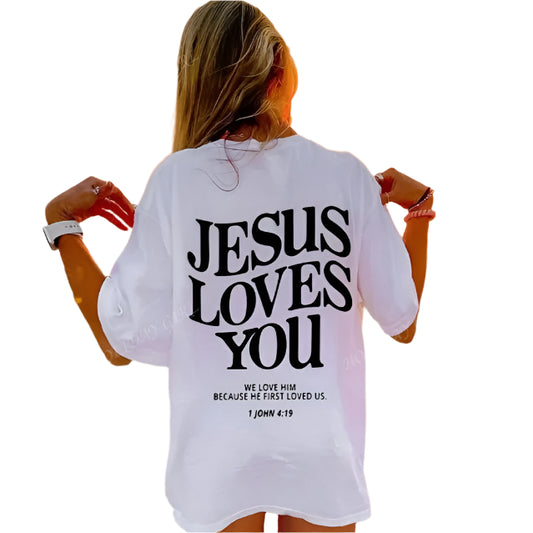 Person wearing a white shirt with 'Jesus Loves You' text on a beach at sunset.