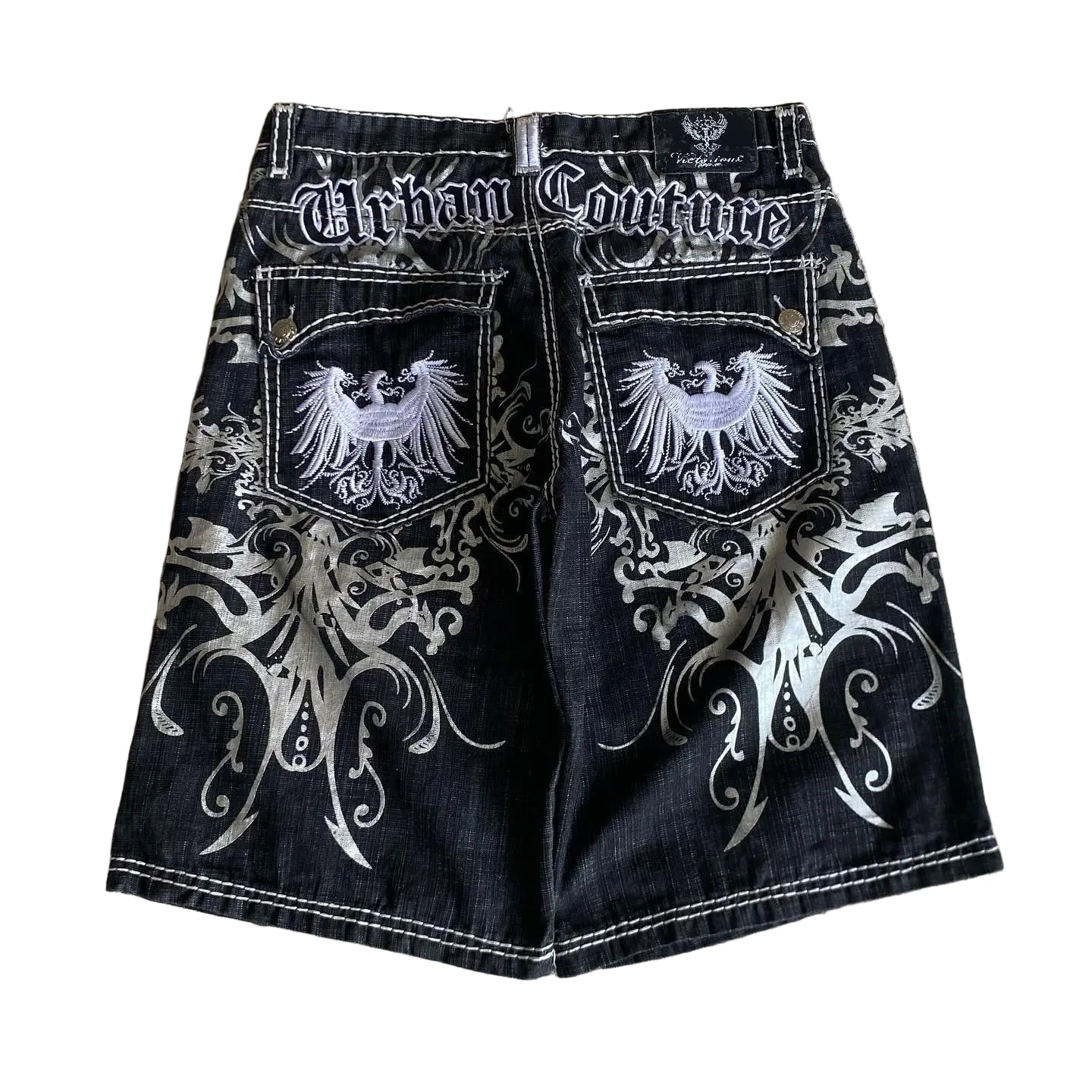 Black denim shorts with white embroidery and 'Urban Couture' branding on a textured wall background.