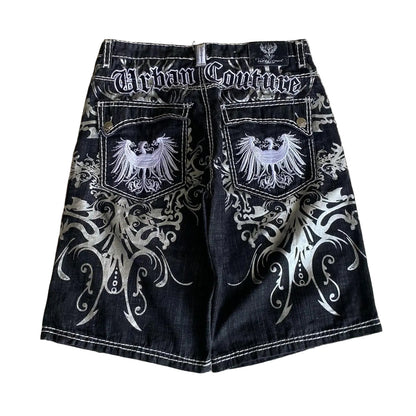 Black denim shorts with white embroidery and 'Urban Couture' branding on a textured wall background.