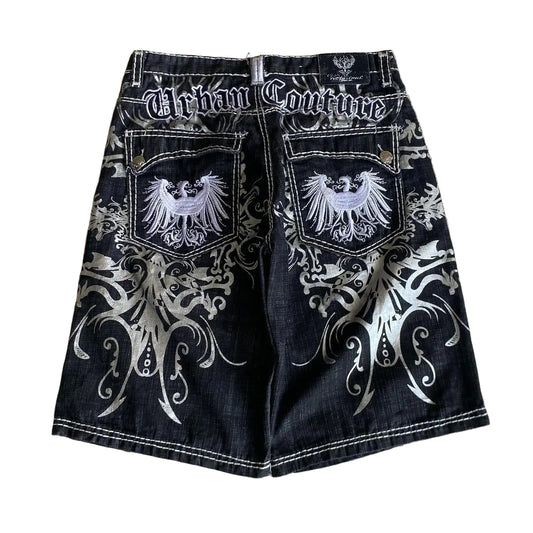 Black denim shorts with white embroidery and 'Urban Couture' branding on a textured wall background.