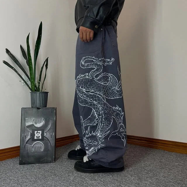 Person wearing dark pants with a white dragon design, standing in a room with a plant and a small table.