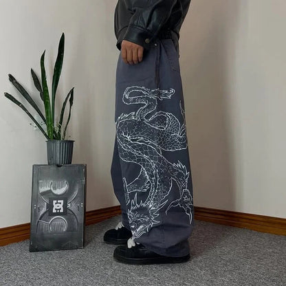 Person wearing dark pants with a white dragon design, standing in a room with a plant and a small table.