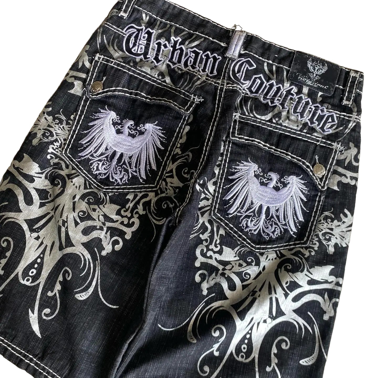Black denim shorts with white embroidery and 'Urban Couture' branding on a beige surface.