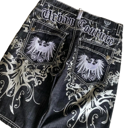 Black denim shorts with white embroidery and 'Urban Couture' branding on a beige surface.