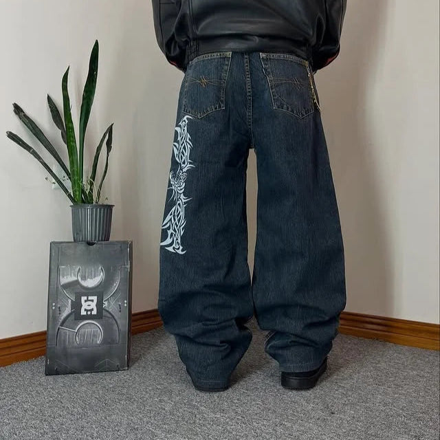Person wearing a black hoodie and dark jeans with white designs, standing in a room with a plant and decorative box.