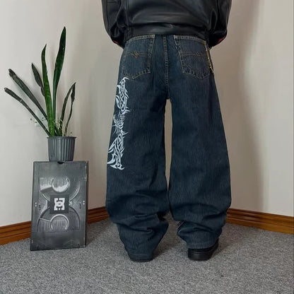 Person wearing a black hoodie and dark jeans with white designs, standing in a room with a plant and decorative box.