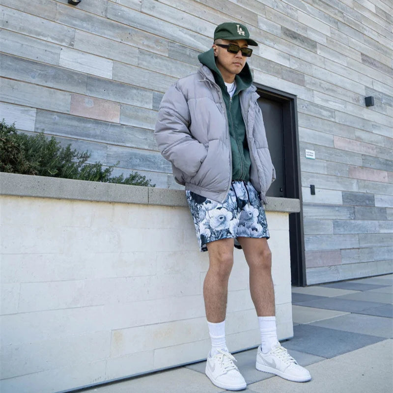 Person wearing a gray puffer jacket, green sweater, floral shorts, and white sneakers standing against a textured wall.